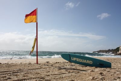 A lifeguard's rescue board sits beside a red and yellow flag flying from a pole on the beach. Attached to the pole is a lifesaving flotation tube. 