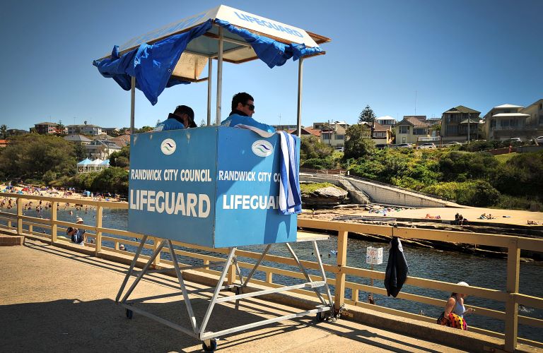 Two lifeguards in blue uniforms and sunglasses sit in a blue lifeguard tower overlooking the water at Clovelly Beach on a sunny day. The water is calm, the sky clear, and writing on the side of the tower reads, Randwick City Council Lifeguards. 