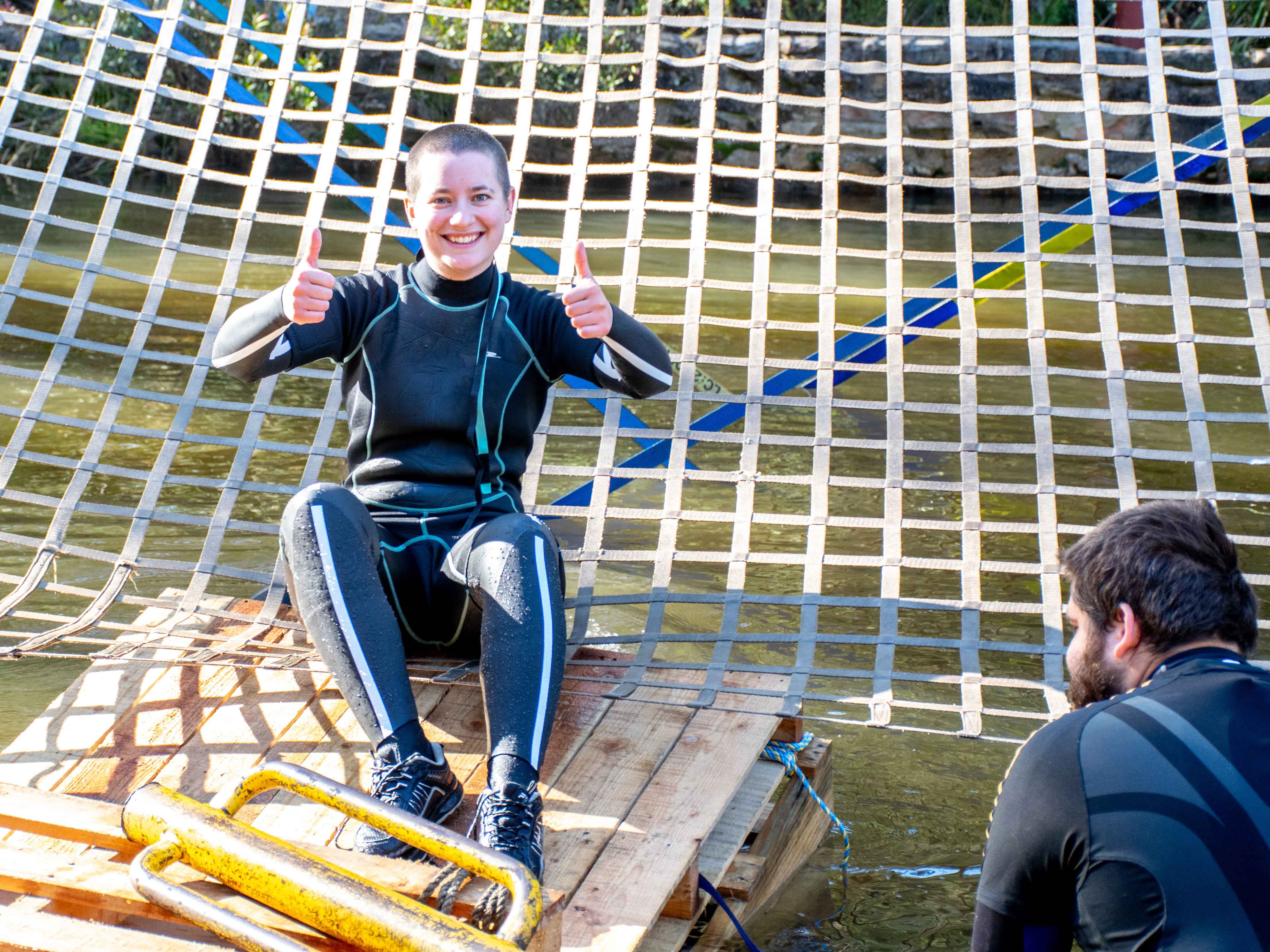 A woman smiling at the camera with her thumbs up and wearing a wetsuit and sitting on a net over water.