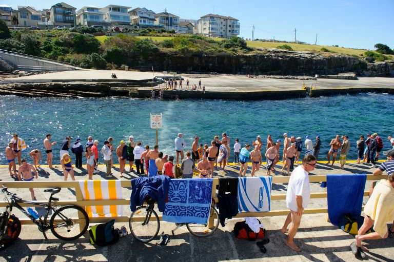 A crowd of men and women wearing swimmers stand on the concrete beside the water at Clovelly Beach. 