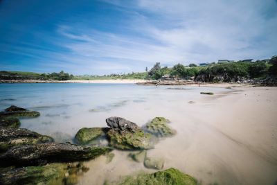 Image of Little Bay Beach on a sunny day. Water is peaceful and blue and a few streaky clouds in the sky. Rocks in the foreground and the green headland in the distance.