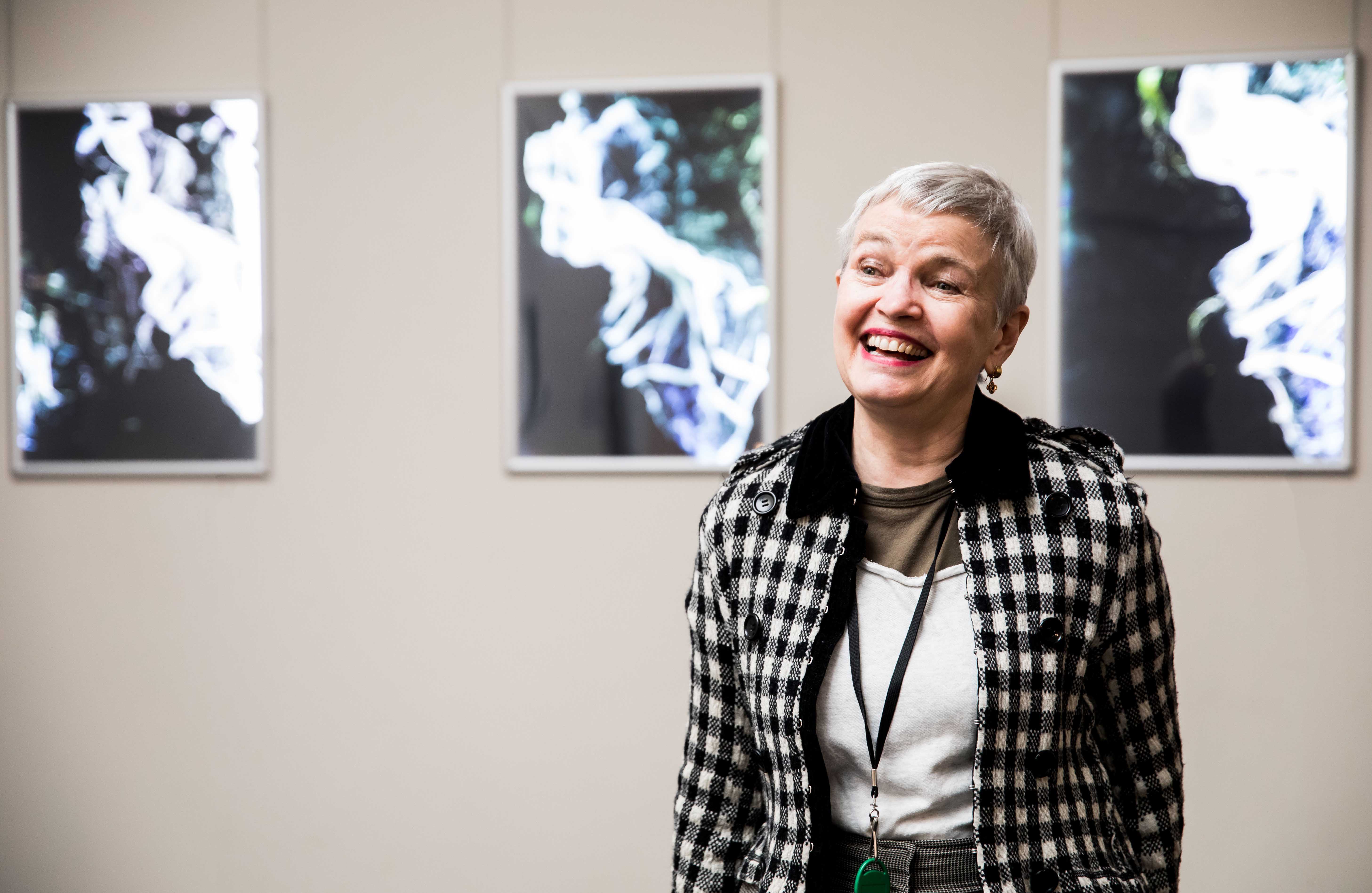 Artist, April Mountfort stands in front of three photographic works mounted on a wall. She has short white hair and smiles to someone off camera.